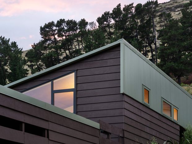 A contemporary hillside dwelling with a steep asymmetrical gabled roof form, dark horizontal cladding, pale corrugated metal upper wall panels, and simple dark-framed windows reflecting the evening sky. Dense trees and a rocky sloping site surround the building.