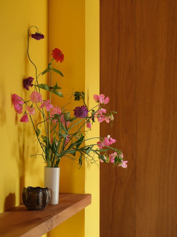 A close-up interior vignette featuring a sculptural floral arrangement in a white cylindrical vase and dark textured vessel, displayed on a thick timber shelf. The backdrop combines a vivid mustard-yellow painted wall with a rich vertical timber door or wall panel, creating a warm, artistic composition with strong colour contrast.