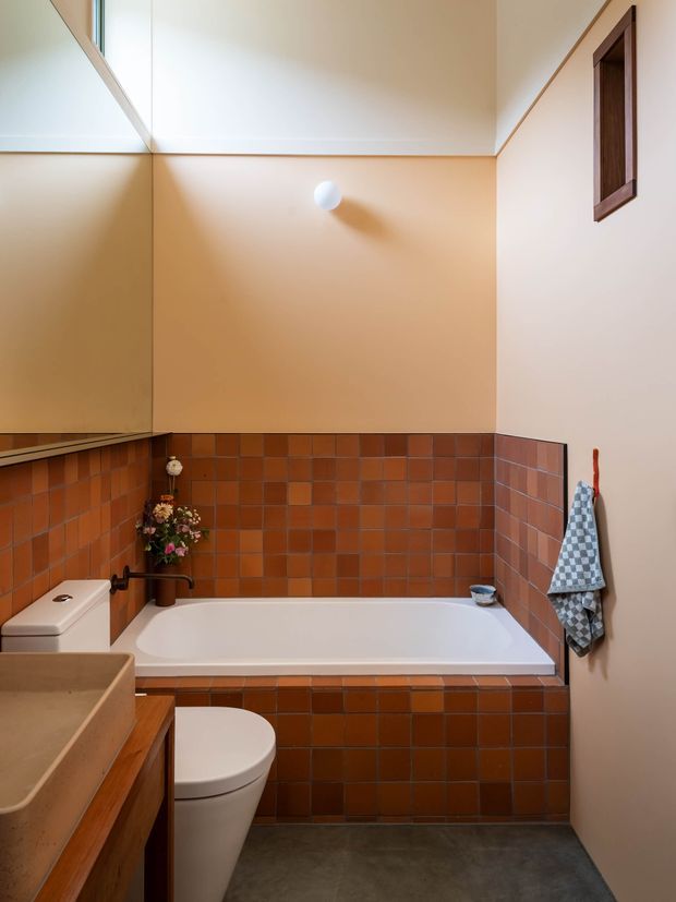 Compact bathroom with a white bathtub set into a raised surround clad in small terracotta-brown square tiles. A matching tiled half-height wall continues beside the tub, while warm peach painted walls rise to a white ceiling. The room includes a white toilet, a timber-toned vanity with a beige basin, a large mirrored wall, dark grey floor finish, and a small framed window. A checked towel and floral arrangement add colour and homely character.