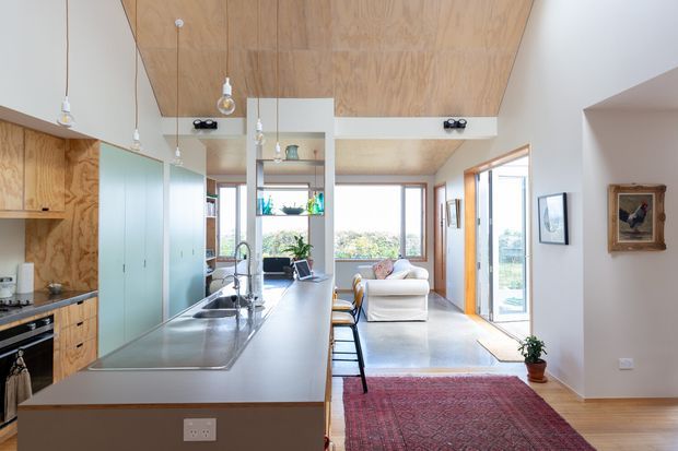 Open-plan kitchen and living area with a long central island, pale timber cabinetry and ceiling panelling, integrated appliances, pendant lights, and expansive glazing. The living zone includes a light sofa, timber flooring, and a large patterned rug. The space is bright and contemporary with warm natural materials.