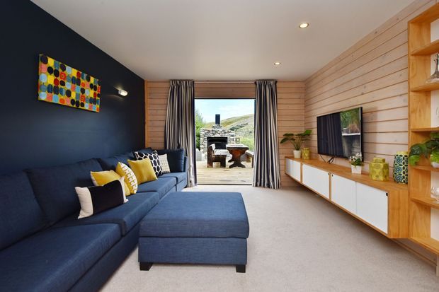 A contemporary living room with a large navy sectional sofa and ottoman, dark blue feature wall, pale timber wall panelling, built-in timber shelving and white-fronted media cabinetry. Full-height striped curtains frame glazed doors opening to an outdoor terrace, with a fireplace visible beyond. The room has recessed ceiling lights, warm timber detailing, and colourful cushions and artwork.