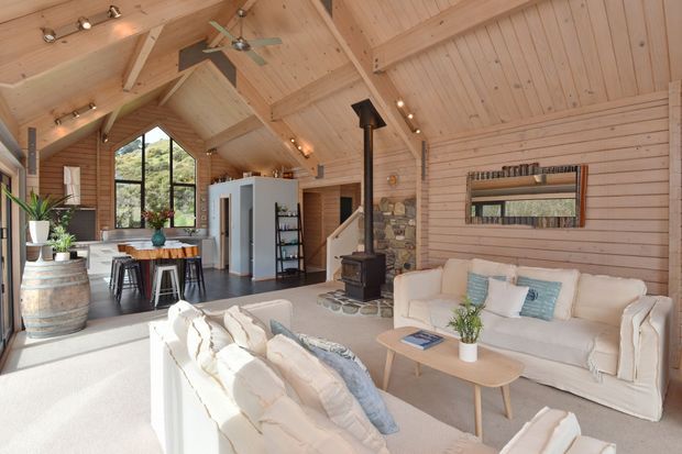 Bright, open-plan living room with a vaulted timber-lined ceiling and matching timber wall panelling. The space includes a white sofa arrangement, pale rug, light wood coffee table, freestanding wood-burning stove with stone surround, compact kitchen or bar area with stools, exposed structural beams, ceiling fans, and large arched glazing overlooking greenery. The room has a warm rustic cabin-like character with contemporary furnishings.