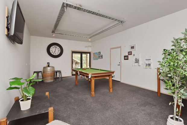 A bright recreational room centered around a full-size green-felt pool table, with dark carpet, white walls, a wall-mounted television, large clock, framed pictures, potted plants, and an exposed rectangular ceiling-mounted lighting frame. A small window and entry door are visible.