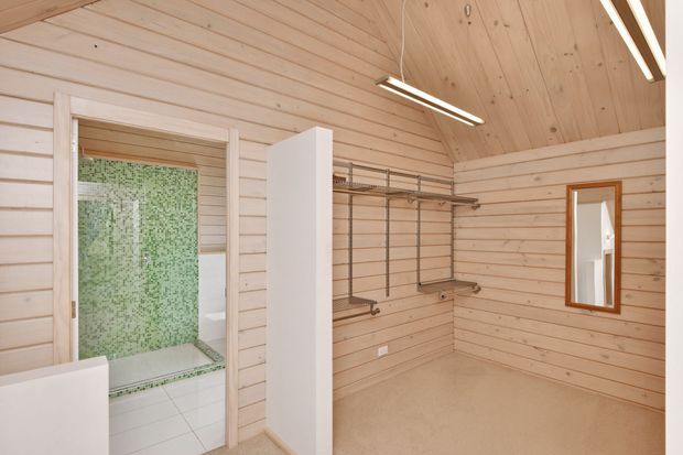 Light-filled timber-lined dressing room or walk-in wardrobe with exposed sloping timber ceiling, open metal clothing rails and shelving, a tall white storage partition, and a small timber-framed mirror. An open doorway reveals an adjoining bathroom with pale green mosaic tile walls and a tiled floor. Slim linear ceiling lights provide contemporary illumination.