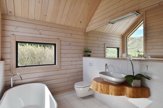 Bright compact bathroom with extensive light timber wall and ceiling panelling, a sloped ceiling, white freestanding bathtub, wall-mounted toilet, vessel basin, and a sculptural live-edge timber vanity shelf. Windows provide views toward a natural hillside landscape.