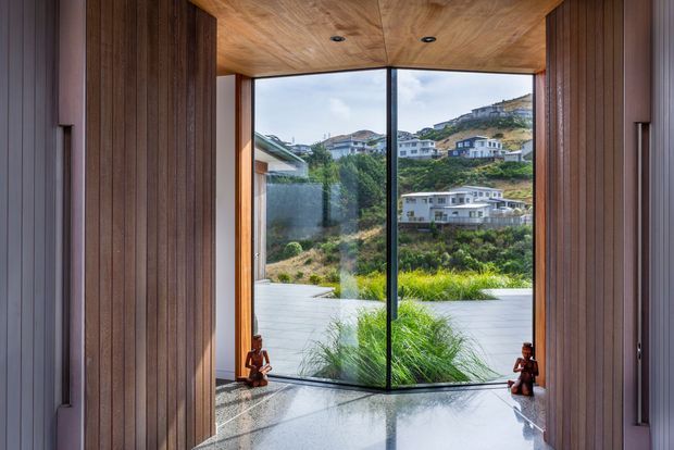 A refined interior threshold or entryway framed by dark vertical timber wall panelling and a warm timber-lined ceiling. Full-height corner glazing opens the view toward a landscaped terrace and a steep, built-up hillside beyond. The polished light stone floor reflects the glazing, while small decorative objects sit at the base of the windows. The composition is minimalist and strongly focused on the framed landscape view.