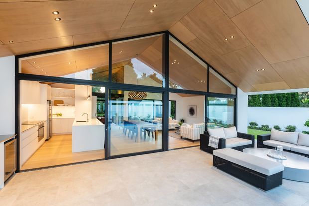 A contemporary indoor-outdoor living area with a dramatic pitched timber-lined ceiling and extensive black-framed glazing. The open-plan space includes a white kitchen with island, dining area, and furnished lounge opening onto a landscaped courtyard. The warm timber ceiling and floors contrast with pale surfaces, dark window frames, and neutral upholstered furniture.