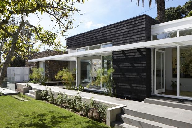 Exterior view of a contemporary single-storey residence with a strong horizontal composition. White rendered or painted structural elements and broad glazing surround a dark shingle-clad upper wall volume. A paved terrace, broad entry steps, low concrete garden walls, manicured lawn, and tropical planting define the front garden. The image is captured in bright daylight.
