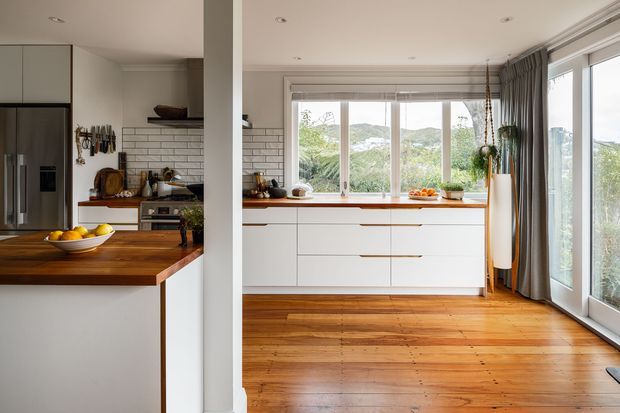 A bright, open-plan kitchen with white flat-panel cabinetry, a warm timber island and timber flooring. The kitchen features a stainless-steel refrigerator, white brick-pattern tiled splashback, timber benchtops, pendant and recessed lighting, and large windows and glazed doors overlooking greenery. Grey curtains frame the glazing, while a central structural column separates the kitchen work area from the adjacent open space.