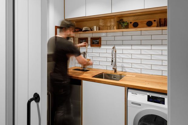 Compact contemporary utility kitchen/laundry space with white flat-panel cabinetry, a warm timber benchtop and open shelf, white subway-tile splashback, stainless-steel sink and tapware, and an integrated front-loading washing machine. A person is blurred in motion at the work surface, and a black-framed glazed door is visible at left.