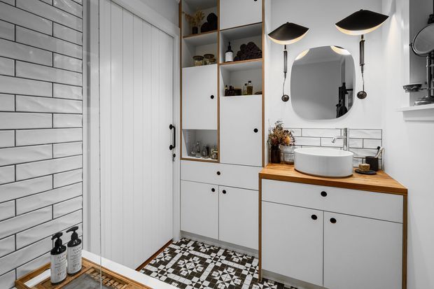 A compact, stylish bathroom with white subway-tile walls, a patterned geometric tile floor, white shaker-style built-in cabinetry, and warm timber benchtops. The vanity features a white vessel basin, rounded mirror, and distinctive black pendant wall lights. A white vertical-panel door and open shelving with toiletries and decor complete the space.