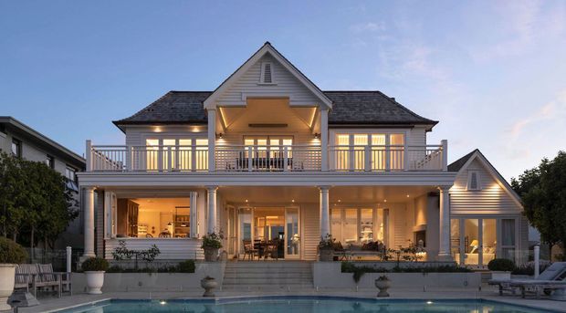 Large two-storey coastal-style residence viewed from the pool, featuring a symmetrical gabled facade, pale horizontal weatherboard cladding, dark shingle roofing, broad balconies, white columns, and warmly lit indoor and outdoor living areas. A swimming pool occupies the foreground.