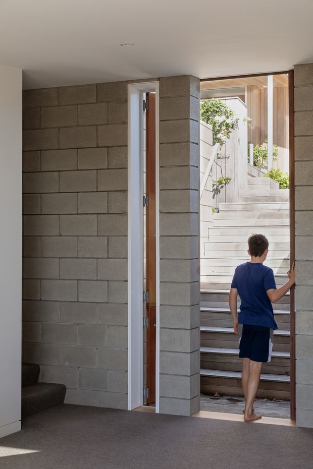 Interior view of a compact stair entry space with exposed grey concrete block walls, dark carpeted floor, and a large glazed opening leading to timber stairs and a bright courtyard-like exterior. A child is shown ascending the stairs. The space has simple modern detailing, recessed ceiling lighting, and timber visible around the stair opening.
