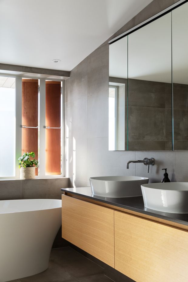 A bright, contemporary bathroom with a freestanding white bathtub, double white vessel basins, a floating light-wood vanity with a dark benchtop, large mirrored cabinetry, grey concrete-look wall and floor tiles, and a narrow vertical window with warm timber shutters. The space has clean lines, abundant natural light, and restrained modern detailing.