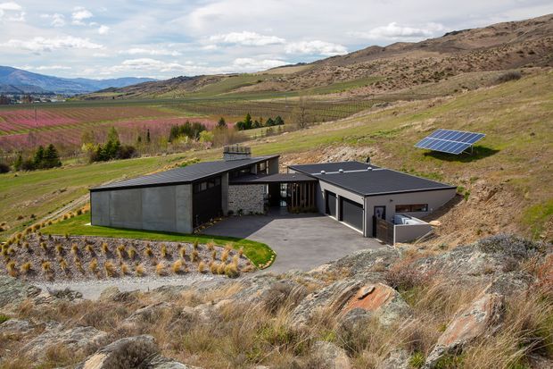 A low-profile modern home set into a dry, sloping hillside, viewed from above. The house has dark flat or very low-pitched roof planes, angular interconnected volumes, dark window and door frames, and a central courtyard-like paved entry. A freestanding solar panel array is visible on the hillside, with cultivated fields and rugged rolling terrain extending behind the property.