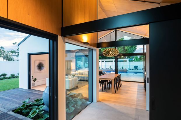 A contemporary indoor-outdoor living and dining area with large black-framed glazed sliding doors opening onto a timber deck and landscaped courtyard. The interior features warm timber ceiling and wall panels, pale flooring, a timber dining table, upholstered seating, and a woven pendant light. Strong horizontal and vertical structural framing creates a connection between the bright interior and outdoor pool or courtyard area.