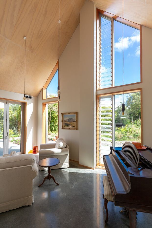 A bright, double-height living room with a polished concrete floor, warm plywood-clad sloping ceiling, tall timber-framed windows and louvred glazing. The room contains light upholstered armchairs, a small round timber coffee table, pendant lights, and a dark upright piano with a chair along the right side. Lush garden views are visible through the glazing.