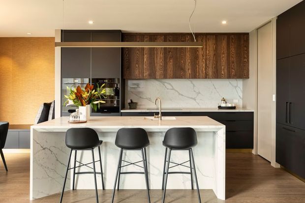 A refined contemporary kitchen with a large white marble-look island and integrated seating for three. The room features dark timber overhead cabinetry, matte black cabinetry and appliances, a white veined stone splashback, warm timber flooring, and a linear pendant light. A dining or living area is partially visible to the left.