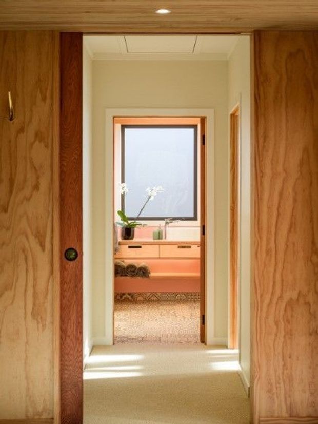 View through a timber-lined hallway toward a softly lit bedroom or dressing area. The foreground features warm wood wall and sliding door panels, carpeted flooring, and a central doorway with contrasting timber trim. Beyond it is a compact room with a large frosted window, low built-in timber cabinetry, a cushioned bench or bed-like surface, and a potted orchid. The overall design is warm, understated, and mid-century influenced.