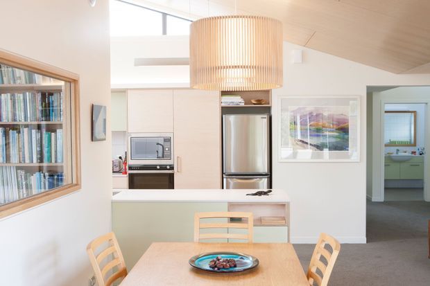 Bright open-plan kitchen and dining area with pale cabinetry, stainless-steel refrigerator, built-in microwave and oven, white island bench, and a light timber dining table with matching chairs. A large slatted timber pendant hangs above the island. Built-in bookshelves are visible through an interior opening on the left, while a hallway leads to a green-toned room on the right. The space has a pitched timber-lined ceiling, clerestory glazing, and a calm, light-filled atmosphere.