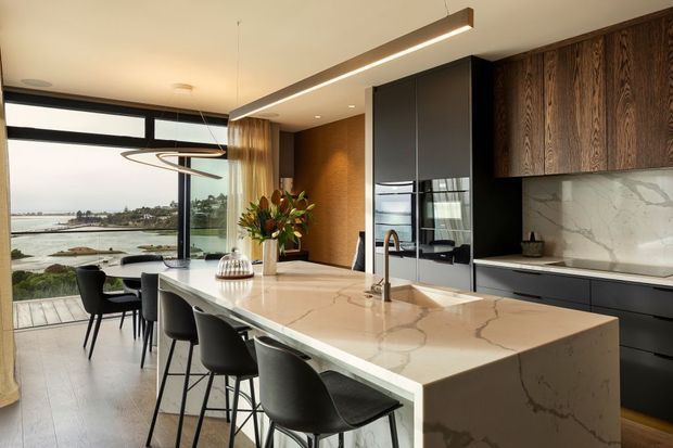 A contemporary kitchen with a large white veined stone island and integrated sink, surrounded by dark timber cabinetry, matte dark appliances, and a marble-look splashback. Black bar stools line the island, while floor-to-ceiling glazing opens to a waterfront outlook and dining area. A linear pendant light runs above the island.