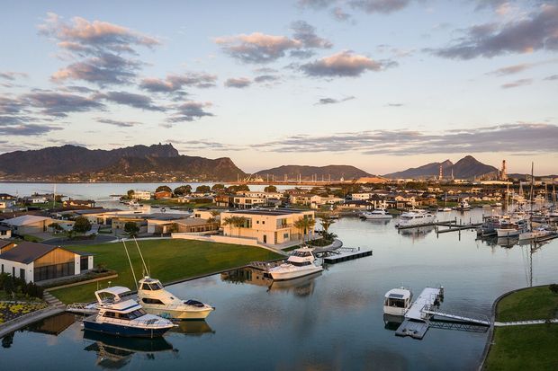 A wide elevated view over a marina-side residential development, with calm waterways, private jetties, moored boats, low-rise homes, landscaped lawns, and a dramatic mountainous backdrop beneath a partly cloudy sky. The scene is presented in warm evening light and has a polished coastal waterfront character.