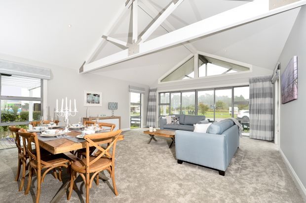 Bright open-plan living and dining room with a vaulted ceiling, exposed white-painted structural beams, large windows, and a triangular clerestory window. The dining area has a timber table with wicker-style chairs, while the living area includes pale blue upholstered sofas, a timber coffee table, and grey patterned curtains. Neutral carpet and light walls create an airy, contemporary coastal feel.