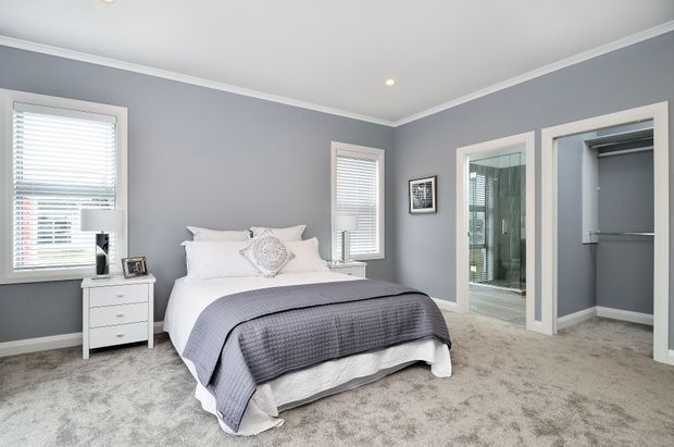 Bright, well-appointed bedroom with a grey upholstered or covered bed, white bedding, grey quilt, white bedside table, grey walls, white trim, carpeted floor, windows with white blinds, and an open walk-in wardrobe. A glass-panelled bathroom is visible through the adjacent doorway.