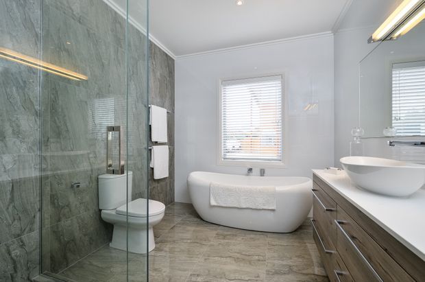 Contemporary bathroom featuring a freestanding white bathtub beneath a window, a glass-enclosed shower with grey stone-look wall tiles, a wall-hung toilet, and a timber-toned vanity with a white vessel basin. The room is bright and clean with mirrored surfaces and recessed lighting.