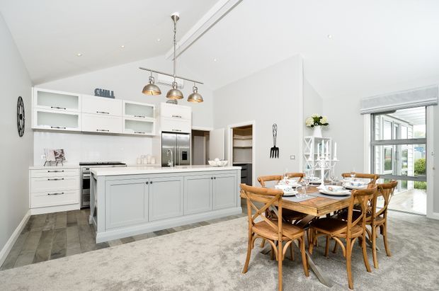 Bright open-plan kitchen and dining area with white shaker-style cabinetry, a large central island, stainless-steel appliances, pendant lighting, and a timber dining table with matching woven chairs. Large windows bring in abundant natural light, while grey-toned flooring and a pale patterned rug create a soft neutral palette.