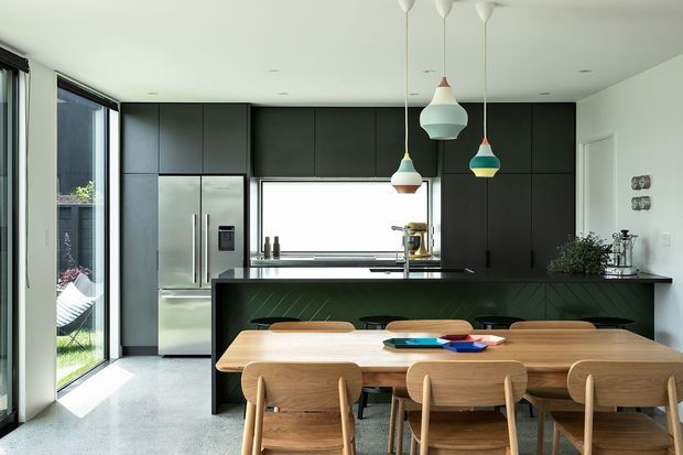 A bright, open-plan contemporary kitchen and dining area. Matte black full-height cabinetry contrasts with a stainless-steel refrigerator and pale stone-look flooring. A dark kitchen island with a subtle diagonal timber detail faces a warm timber dining table and matching chairs. Large glazed doors and windows bring in abundant daylight, while colourful pendant lights provide visual accents.