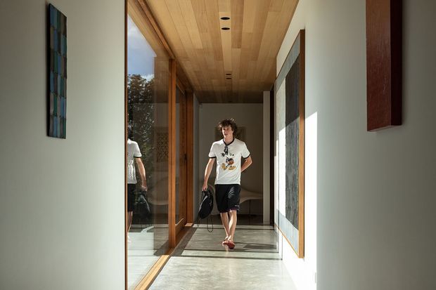 A bright, narrow contemporary hallway or gallery corridor with floor-to-ceiling timber-framed glazing on the left, a warm timber-lined ceiling with recessed downlights, polished concrete flooring, and large wall-mounted artworks. Strong sunlight creates a high-contrast, airy interior, with a person walking through the corridor.