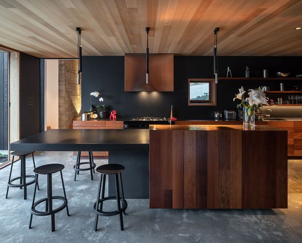 A sophisticated contemporary kitchen with a dark charcoal wall backdrop, a large black island and integrated breakfast bar, and a timber-clad island base. The space features rich dark-wood cabinetry, a warm timber slatted ceiling, black pendant lights, polished concrete flooring, open shelving, and a compact window above the rear bench. Several black stools line the island, creating a dramatic yet warm entertaining-focused kitchen.