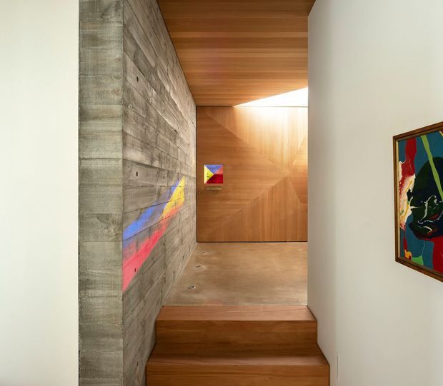 A refined contemporary interior stairway and landing with warm timber lining, a polished concrete wall and floor, and recessed lighting. The compact stair rises beside a textured concrete feature wall, leading to a timber-clad landing with a bright clerestory opening. Colourful abstract artworks provide small accents against the neutral architectural palette.
