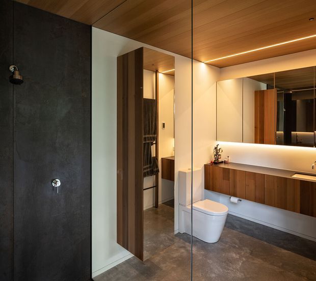 A refined contemporary bathroom featuring a dark textured shower wall enclosed by frameless glass, a wall-mounted timber vanity with a pale benchtop, a large mirrored splashback, warm timber ceiling lining, recessed linear lighting, and a white toilet. A timber door opens toward a partially visible adjoining space. The palette is warm, dark, and minimalist with integrated architectural lighting.
