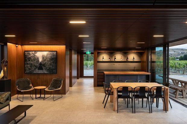 A sophisticated open-plan hospitality or residential entertaining space with a dark timber slatted ceiling, warm timber wall panelling, integrated recessed lighting, and pale stone-look flooring. The room includes a dining table with black chairs, a central timber-topped island or bar, a back bar with bottle display and cabinetry, lounge chairs, and extensive glazing opening toward a landscaped vineyard-like setting. The interior has a warm, refined contemporary character with strong indoor-outdoor connection.