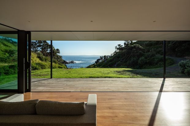 A modern living room opens directly onto a broad timber deck through expansive floor-to-ceiling glazing and sliding doors. The room has a low, plain ceiling, warm timber flooring, and a simple upholstered sofa in the foreground. Outside, a grassy coastal landscape and ocean view are framed by the glazing, with strong afternoon sunlight casting long shadows across the floor and deck.