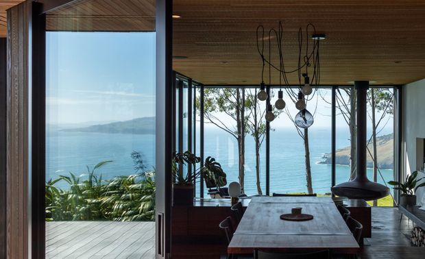 A refined dining area with a long timber dining table and sculptural hanging pendant lights, framed by extensive floor-to-ceiling glazing. The room opens onto a timber deck and overlooks a calm coastal bay, with timber ceilings, dark window frames, and abundant indoor planting creating a strong connection to the landscape.