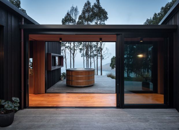 A dark-clad contemporary pavilion or house opens onto a timber deck, framing a freestanding round timber soaking tub and a tranquil wooded waterfront landscape beyond. Large sliding glass doors, warm timber lining, black exterior cladding, and strong indoor-outdoor connection dominate the composition.