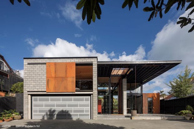 Exterior view of a contemporary multi-level residence with a strong geometric composition. The facade combines light grey masonry blockwork, large orange-rust metal feature panels, extensive dark-framed glazing, and a broad projecting flat roof with an exposed grid-like soffit. A translucent sectional garage door occupies the front, while a covered glazed entry or outdoor living zone sits to the right. The house is photographed from a paved driveway under a bright blue sky, with surrounding planting and neighbouring structures visible.