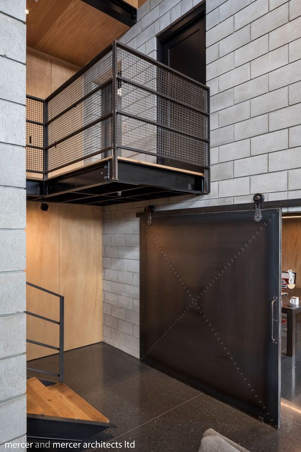 Interior architectural photograph of an industrial-style multi-level space featuring a black steel mesh mezzanine and guardrail, a large dark sliding barn-style door with exposed hardware, grey concrete block walls, warm timber wall and ceiling lining, and a polished dark concrete floor. A stair and partial seating area are visible, with strong contrast between black metal, concrete masonry, and timber finishes.