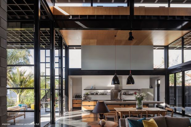 A spacious open-plan contemporary interior combining a kitchen, dining area and living space beneath a dramatic double-height volume. Full-height black-framed glazing surrounds the room, with exposed dark structural beams, a warm timber-lined ceiling, white built-in kitchen cabinetry and a large central island. A timber dining table, pendant lights and living seating occupy the foreground, while tropical planting is visible outside.