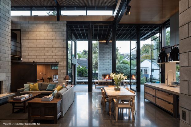 A refined open-plan living, dining and kitchen interior with exposed grey masonry walls, dark steel-framed floor-to-ceiling glazing, polished concrete flooring and a timber-lined ceiling. The room features a large sectional sofa, dark timber coffee table, dining table and chairs, and a streamlined kitchen with timber cabinetry and a light benchtop. An outdoor courtyard and neighbouring residential buildings are visible through the glazing.