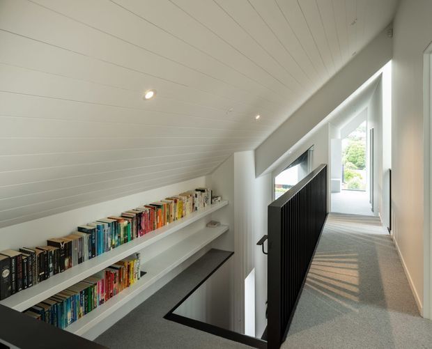 Bright upper-level hallway or landing beneath a sloped, white-lined ceiling. Long built-in white bookshelves filled with books run along the left wall, while a black vertical-slat balustrade guards an open stairwell at centre. Carpeted flooring, recessed lighting, and large windows at the end create a light-filled, contemporary reading and circulation space.