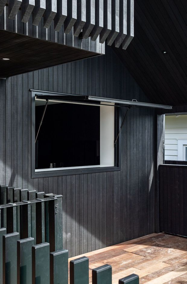 A dark, modern exterior courtyard or terrace clad in vertically grooved charcoal timber or composite boards. The space features a large horizontal awning window, a black slatted pitched roof with projecting exposed beams, a dark vertical-slat balustrade, and warm brown tiled or timber-like paving. A neighbouring light weatherboard facade is visible beyond the boundary.