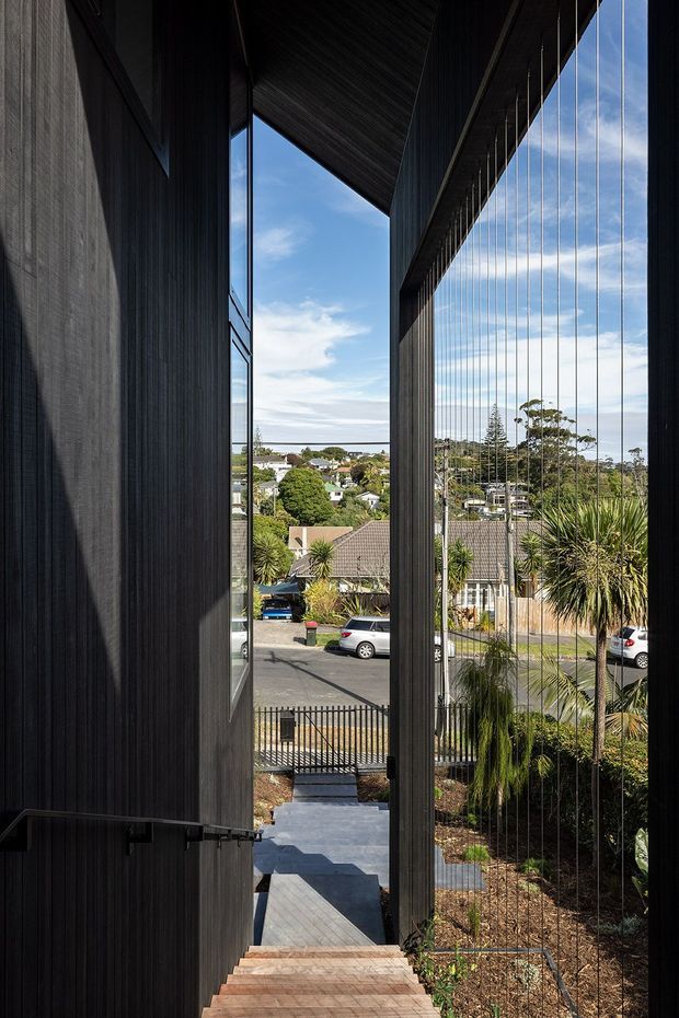 A dramatic architectural view from a narrow stairway or passage, framed by tall dark vertically clad walls and a large sloping roof overhang. Fine vertical cables form a transparent balustrade along the right side, overlooking a landscaped garden, residential street, and suburban houses beyond. The composition emphasises strong vertical lines, deep shadows, natural light, and an elevated outlook.