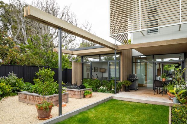 Contemporary residential courtyard and rear facade with a timber-clad upper level, extensive glazing, slim dark structural posts, and a broad timber pergola framing a paved outdoor entertaining area. Raised garden beds, gravel landscaping, lawn, potted plants, and a covered dining space create a landscaped indoor-outdoor connection.