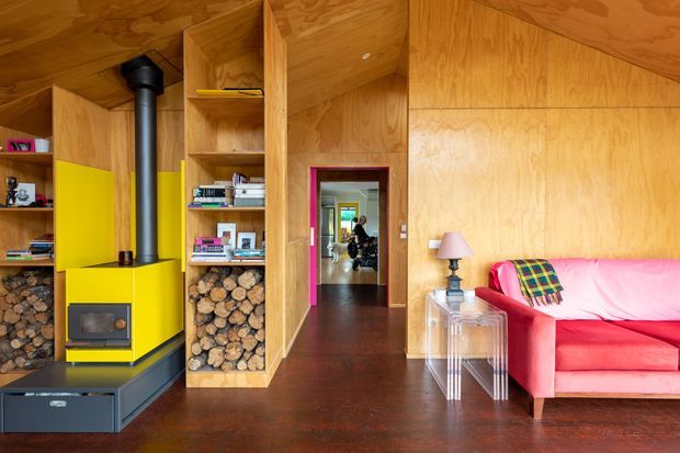 A compact, warm timber-lined living room with extensive built-in plywood cabinetry and shelving. A bright yellow wood-burning stove forms a strong focal point beside neatly stacked firewood and books. The room has dark timber flooring, a vivid pink sofa with a multicoloured throw, a small transparent side table and lamp, and a hallway leading to further rooms.