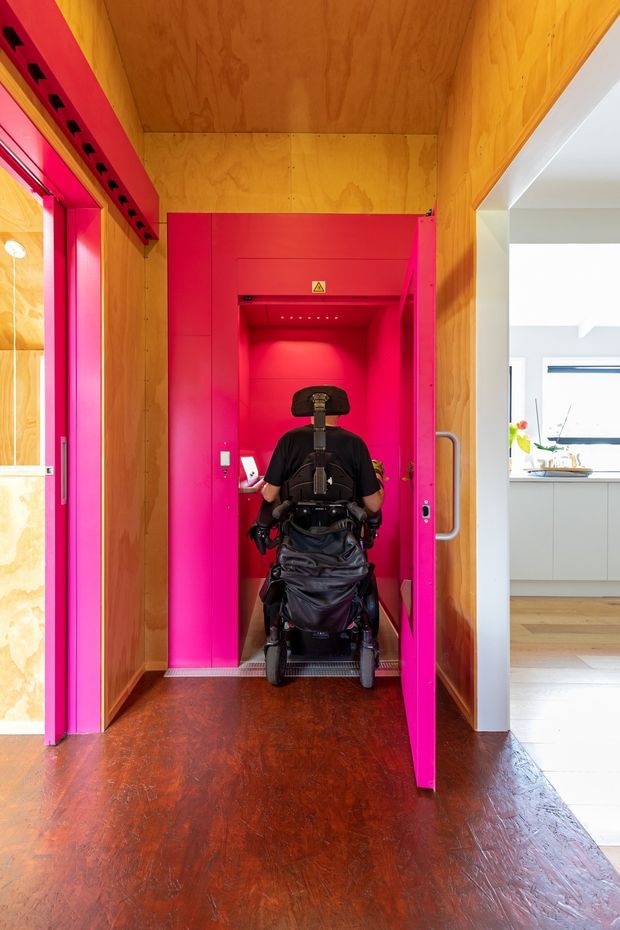 Interior view of a compact residential entryway featuring a vivid hot-pink wheelchair-accessible lift or elevator enclosure. The surrounding walls and ceiling are clad in warm plywood, with a dark reddish-brown timber floor. A person using a wheelchair is entering the lift. A bright white kitchen or living area is visible through the opening on the right.