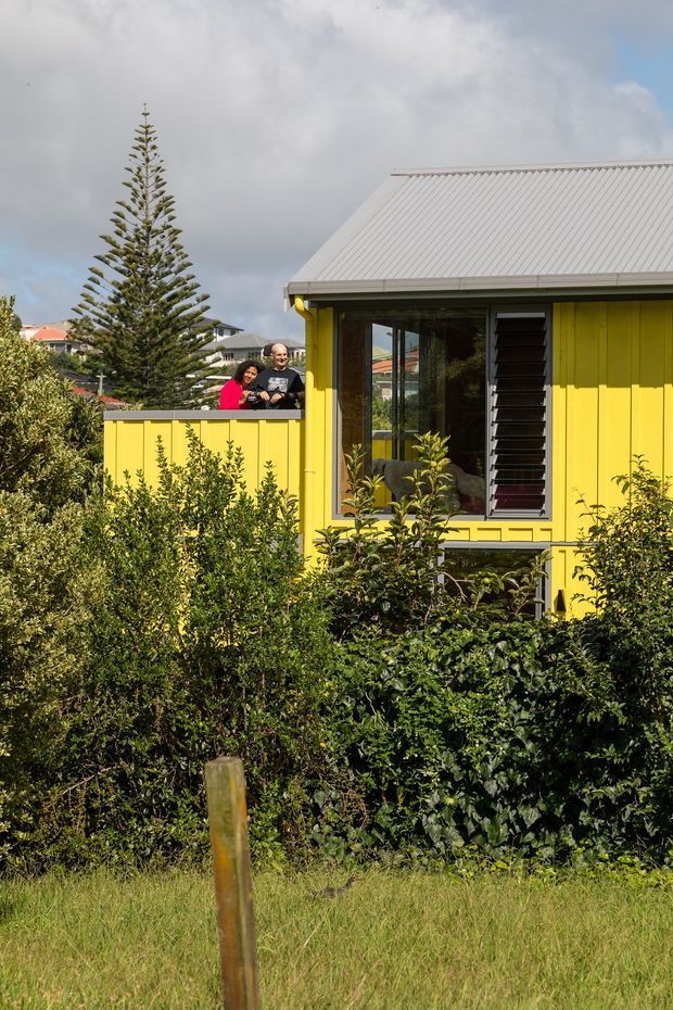 Exterior view of a compact, brightly painted yellow weatherboard house with a light grey corrugated metal gable roof. The facade features dark-framed glazing, a tall narrow louvred window, and simple decorative trim. Dense landscaped planting fills the foreground, with a yellow boundary wall and two people visible on an elevated outdoor area behind it. Other residential roofs and a tall conifer appear in the background.